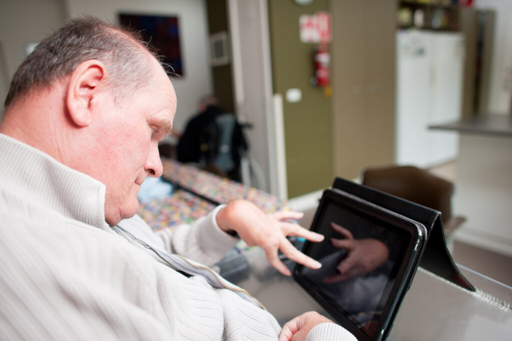 Mature aged man with a disability operating touchscreen computer. The reflection of his hand can be seen on the screen. He is seated in his living room.