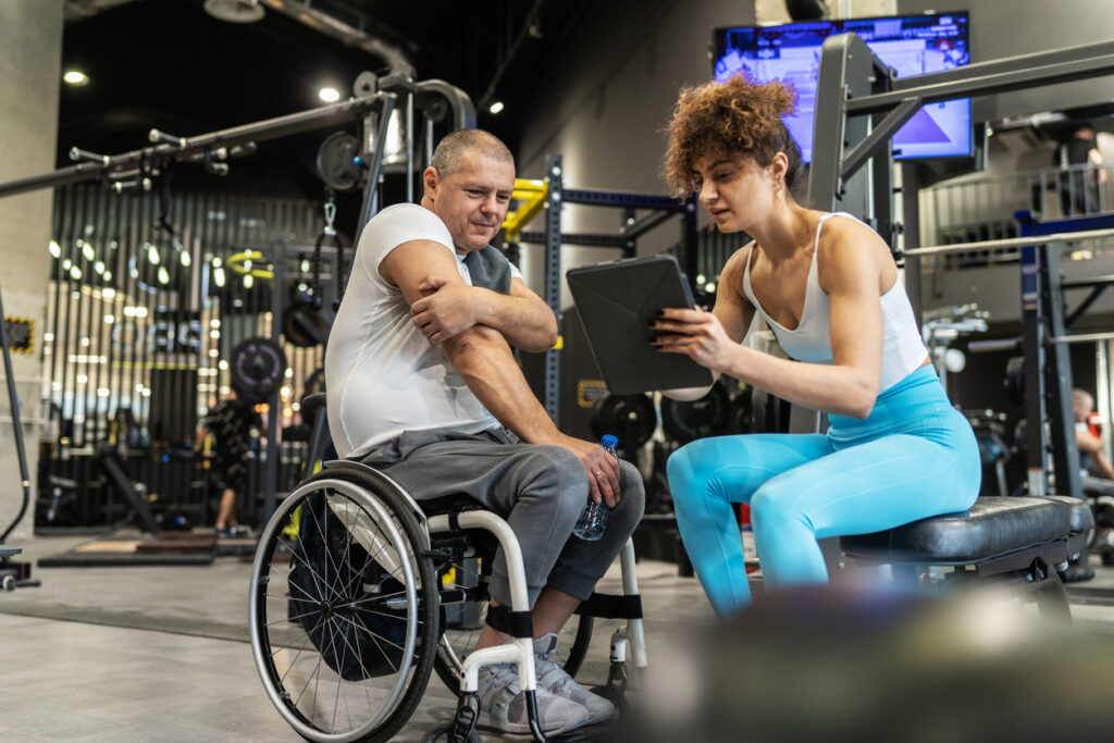 A middle aged man sitting in his wheelchair without obstacles is at the gym with his female instructor. 