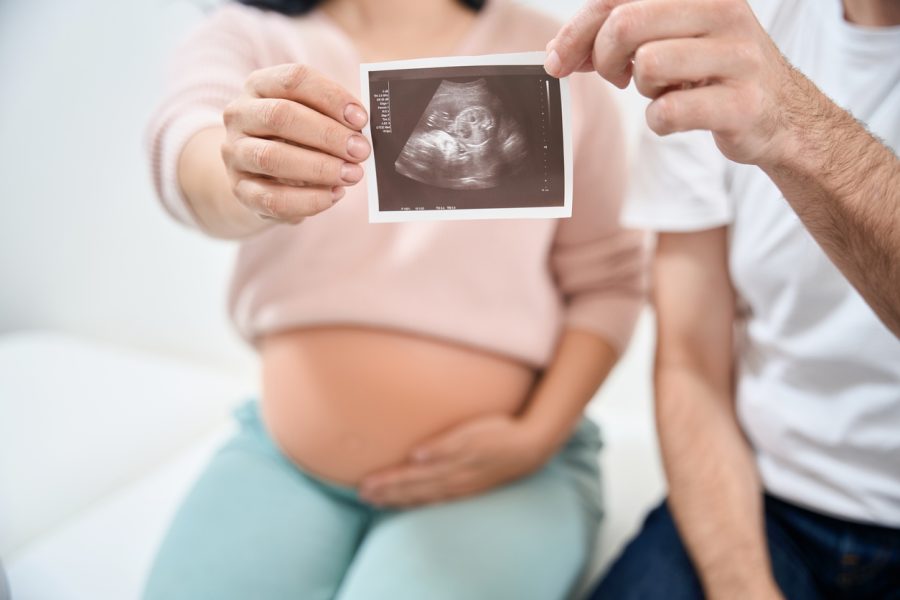 Happy couple sitting on examination couch in doctors office and holding the ultrasonography picture of their healthy child in hands, deep love and affection, healthcare and medicine