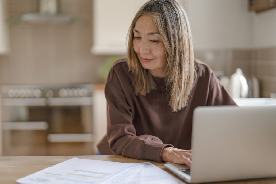 A woman with long hair is engrossed in her laptop, surrounded by scattered papers and documents. She embodies concentration while sitting at a wooden table in a well-lit kitchen.